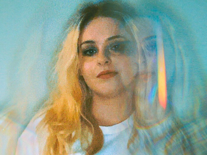 emzae, with blonde hair and a multicoloured blur in the background
